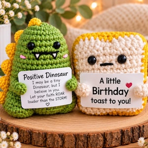 May include: Two crochet plushies: a green dinosaur with yellow spikes and a white-and-yellow toast slice. The dinosaur holds a sign reading Positive Dinosaur, I may be a tiny Dinosaur, but I believe in you. Let your faith ROAR louder than the STORM! The toast holds a sign reading A little Birthday toast to you!