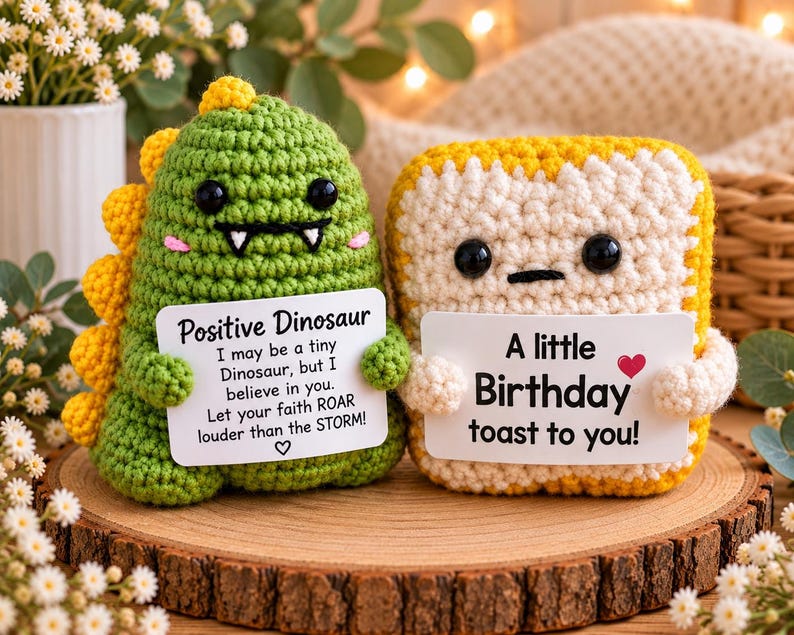 May include: Two crochet plushies: a green dinosaur with yellow spikes and a white-and-yellow toast slice. The dinosaur holds a sign reading Positive Dinosaur, I may be a tiny Dinosaur, but I believe in you. Let your faith ROAR louder than the STORM! The toast holds a sign reading A little Birthday toast to you!