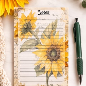 May include: A notepad with a sunflower design and the word "Notes" at the top. The notepad has lined pages and is decorated with watercolor sunflowers and green leaves. A green pen rests on the right side of the notepad.