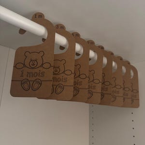 Peut inclure: Ensemble de cintres pour vêtements de bébé en carton marron, avec un ours et le texte "1 mois". Les cintres, marqués des mois, sont suspendus à une tringle blanche dans un placard blanc.