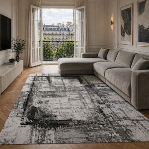 May include: A large rectangular area rug with a distressed abstract design in shades of gray and black. The rug is placed on a hardwood floor in a living room setting, with a gray sectional sofa and a view of a city through open French doors.