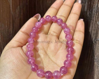 May include: A pink beaded bracelet with the text "Minimagic Jewels" is held in a hand. The bracelet is made of round, polished beads in varying shades of pink. The beads are strung together to form a flexible band.