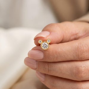 Pu&ograve; includere: Un orecchino a lobo dorato a forma di testa di topo, con una grande gemma rotonda e trasparente al centro e due gemme pi&ugrave; piccole per le orecchie. L'orecchino &egrave; tenuto in mano.
