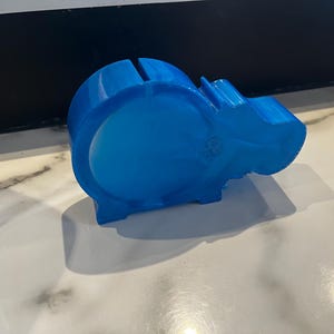 Puede incluir: Un cerdito con forma de hipopótamo azul translúcido. El cerdito tiene una ranura en la parte superior para insertar monedas y un cuerpo redondo. La cabeza del hipopótamo es visible en el lado derecho. El cerdito es de plástico.