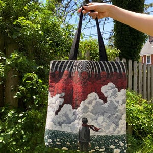May include: A black tote bag with a detailed tapestry design. The artwork depicts a scene with red figures, white clouds, and a person with a red scarf. The bag has black handles and is held up against a backdrop of green foliage.