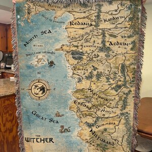 May include: Woven throw blanket featuring a detailed map of a fantasy world, with place names like Redania and Temeria. The map is in shades of blue, tan, and green, with a compass rose and the text "The Witcher". The edges have a fringed border.