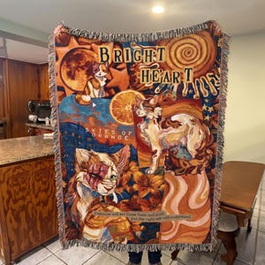Op de afbeelding: Een geweven sprei met een kleurrijk ontwerp met katten, sinaasappels en de woorden "BRIGHT HEART" en "SKIES OF ORANGE". De deken heeft een gefranjerde rand en bevat de tekst: "Everyone will see those flaws and scars, just the right one will understand."