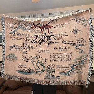 May include: A beige tapestry throw blanket featuring a map of Middle-earth with illustrations of mountains, rivers, and creatures. The map includes text such as "The Lonely Mountain" and "The Desolation of Smaug". The blanket has a fringed edge.