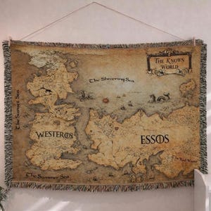 Può includere: Un arazzo raffigurante una mappa del mondo conosciuto, con i testi "The Known World", "Westeros" e "Essos". La mappa è nei toni del marrone e beige, con un bordo a frange. L'arazzo è appeso a una parete.