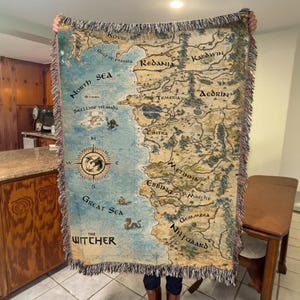 May include: A woven throw blanket with a map design, featuring the text "The Witcher." The map shows regions such as Redania, Temeria, and Nilfgaard, in blue and tan. The blanket has a fringed edge, and is likely made of cotton or a similar material.