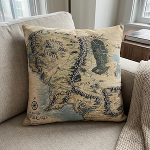 Könnte beinhalten: Beiges quadratisches Kissen mit einer detaillierten Karte von Mittelerde, einschließlich Regionen wie Rohan, Gondor und Mordor. Das Kissen hat eine Vintage-Ästhetik mit Textbeschriftungen und einer Windrose. Es liegt auf einem hellen Sofa.