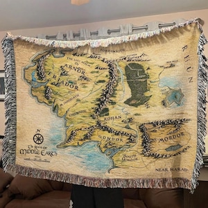 Puede incluir: Una manta beige con un mapa de la Tierra Media, con nombres de lugares como Gondor y Mordor. La manta tiene un borde con flecos y el texto "The Realm of Middle Earth". El mapa muestra detalles geográficos.