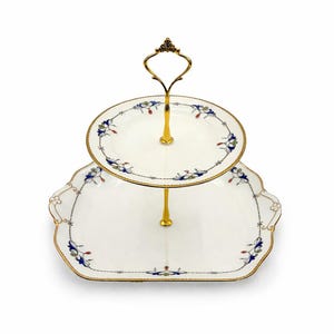 May include: Two-tiered serving tray with a gold-colored handle and supports. The white porcelain plates have a scalloped edge and are decorated with blue and gold floral patterns. The tray is ideal for serving desserts or appetizers.