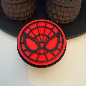 May include: A round, red and black Spider-Man themed coaster. The coaster features a black web design over a red background, with a black border. The coaster is sitting on a black surface.