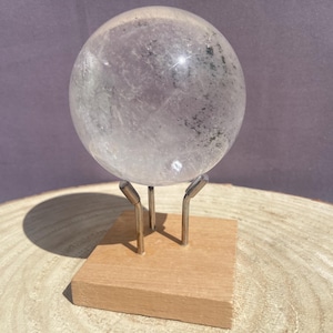 Peut inclure: Une boule de cristal de quartz sphérique et transparente avec des inclusions et des fractures internes visibles. La sphère est soutenue par un support en métal argenté à trois branches fixé sur une base carrée en bois clair.
