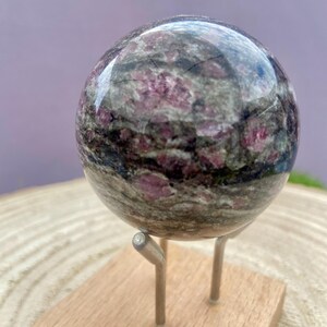 Peut inclure: Une sphère de pierre précieuse polie, principalement grise avec des veines rose foncé et noires, est présentée sur un petit socle en bois avec des supports métalliques argentés. La surface de la sphère reflète la lumière.