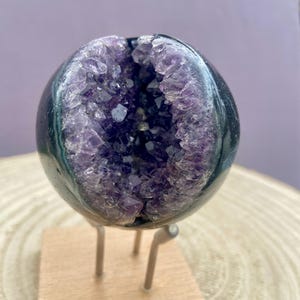 Peut inclure: Une sphère polie et sombre avec une cavité remplie de cristaux d'améthyste violets. La sphère est montée sur une petite base en bois avec des supports métalliques, mettant en valeur la beauté naturelle de la pierre précieuse. Géode d'améthyste.
