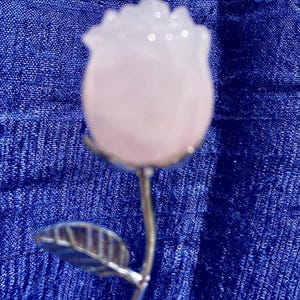Peut inclure: Une sculpture de rose décorative composée d'une fleur en pierre translucide rose pâle fixée sur une tige métallique fine et polie de couleur argentée. La tige présente deux petites feuilles métalliques détaillées sur les côtés.