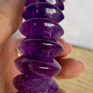 Peut inclure: Un bracelet composé de multiples perles d'améthyste violet foncé polies. Chaque perle présente une forme de disque aplati distincte avec des bords arrondis, créant un aspect empilé uniforme. Les pierres violettes translucides reflètent la lumière.