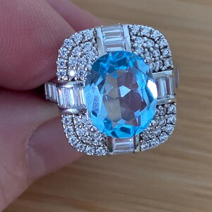 Peut inclure: Une bague en argent ornée d'une grande pierre précieuse ovale bleu clair en son centre. La bague est entourée de petits diamants transparents et d'éléments rectangulaires, créant un design étincelant et élégant. La bague est tenue par une main.