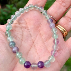 Peut inclure: Un bracelet de perles composé de pierres lisses, rondes et translucides dans des tons variés de vert clair, transparent et violet profond. Les perles sont enfilées sur un cordon fin et flexible, formant un cercle à la finition polie et brillante.