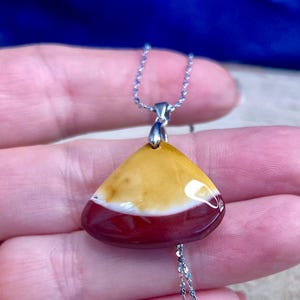 Peut inclure: Un collier pendentif avec une pierre triangulaire polie. La pierre présente un dégradé de couleurs jaune, blanc et rouge foncé. Le pendentif est suspendu à une fine chaîne argentée, créant un accessoire simple mais élégant.