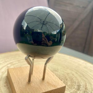 Pode incluir: Uma esfera preta polida, refletindo um guarda-chuva, é exibida em uma pequena base de madeira com três pernas de metal. A superfície da esfera é altamente reflexiva, criando um efeito de espelho. A base de madeira é marrom claro.