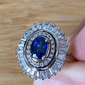 Peut inclure: Une bague en argent ornée d'une grosse pierre précieuse ovale bleu foncé au centre, entourée de petites pierres claires rectangulaires et rondes. La bague a un design en halo avec plusieurs couches de pierres scintillantes.