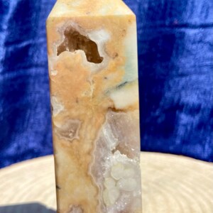 Op de afbeelding: Een gepolijste kristallen toren met een puntige bovenkant, voorzien van een gemarmerd patroon in warme beige, crème en zachte oranje tinten. Het stenen oppervlak vertoont natuurlijke kristallijne insluitingen en een kleine, holle opening nabij de top.