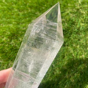 Peut inclure: Une pointe de cristal de quartz clair avec une pointe facettée. Le cristal est transparent avec des stries internes et se trouve sur un fond d'herbe verte. Le cristal est tenu dans une main.