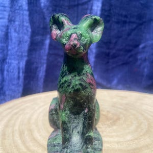 May include: A carved stone cat figurine in shades of green and pink. The cat is sitting upright with its ears perked up. The stone has a speckled appearance and is set on a wooden surface.