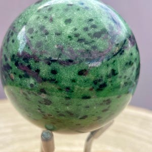 Peut inclure: Une sphère verte polie avec des taches sombres et des stries violettes. La pierre précieuse est présentée sur un support en métal argenté. Il s'agit d'un objet décoratif, peut-être pour l'exposition ou la méditation.