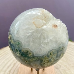 Peut inclure: Une sphère en agate polie avec un intérieur de type géode. La sphère présente un mélange de teintes claires, blanches et vertes, avec une ouverture rugueuse et cristalline. Elle est présentée sur un petit socle en bois avec des pieds en laiton.