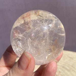 Op de afbeelding: Een heldere, bolvormige kwartskristal met interne breuken, natuurlijke insluitingen en subtiele roodbruine mineraalafzettingen door de transparante steen, met een gepolijst, glad oppervlak dat het licht reflecteert.
