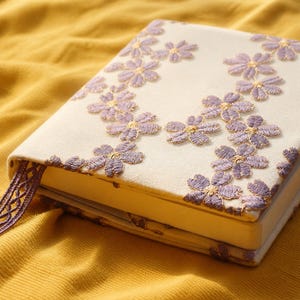 May include: A hardcover book featuring a cream-colored fabric cover embroidered with a pattern of small purple flowers. The book includes a matching purple woven ribbon bookmark extending from the side, with the floral embroidery continuing along the edges.