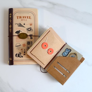 May include: A tan travel notebook with a dark brown spine and a cover featuring illustrations of an umbrella, a bird, and a camera with the text TRAVEL NOTES. Beside it, an open brown paper folder with two orange circular fasteners and slots.