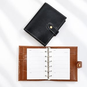 May include: A black leather personal planner with white stitching and a gold snap closure, positioned above an open brown leather planner featuring a six-ring metal binder mechanism and white paper inserts with printed daily task lists.