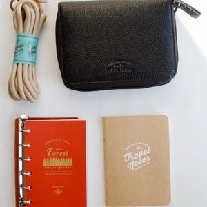 May include: A black pebbled leather zippered case, a beige cord with a blue label, a red loose-leaf notebook with gold text reading Forest, and a tan notebook with white text reading Travel Notes. The items are arranged on a white surface.