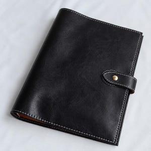 May include: A black notebook with a textured surface and white contrast stitching along the edges. The cover features a secure strap closure with a single gold-toned metal snap button, keeping the notebook closed and protected.