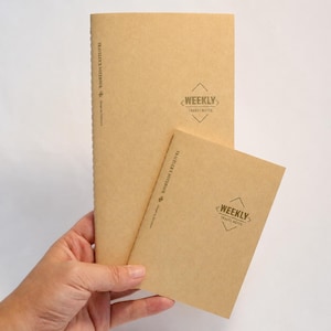 May include: Two tan paper notebooks with stitched spines. The larger notebook features the text TRAVELER'S NOTEBOOK and a diamond-shaped logo with the text WEEKLY TRAVEL NOTES. The smaller notebook displays the same text and logo design.
