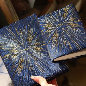May include: Two rectangular journals with textured covers featuring a dark blue base with raised, radiating lines in metallic gold and silver. The covers have a dense, starburst-like pattern that extends from the center toward the edges.