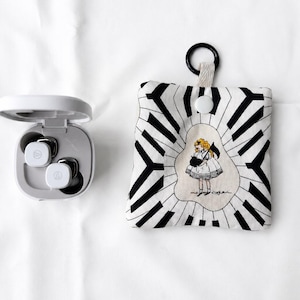 May include: A white wireless earbud charging case with two white earbuds inside, positioned next to a square fabric pouch featuring a black and white piano key pattern and a central illustration of a girl holding a black cat with a white dress.