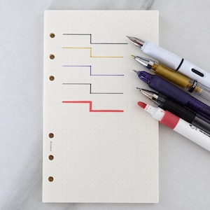 May include: A cream-colored dot grid planner refill page with six punched holes on the left side and the text L-100n printed near the bottom. Five horizontal step-pattern lines in black, gold, purple, black, and red are drawn next to five pens.