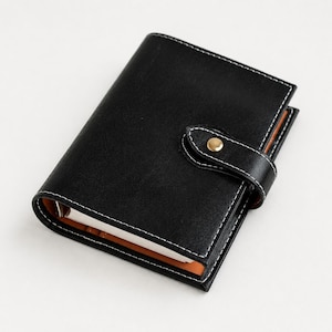 May include: A black leather-like personal planner with white contrast stitching. The cover features a secure strap with a gold-toned metal snap button closure. The interior reveals orange lining and white paper pages held by a metal ring mechanism.