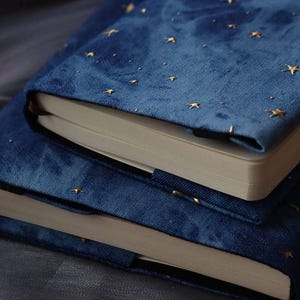 May include: Two stacked notebooks featuring deep blue velvet covers adorned with small, embroidered gold stars. Each notebook has a thick, rectangular shape with cream-coloured pages visible along the edges and a black elastic band closure.