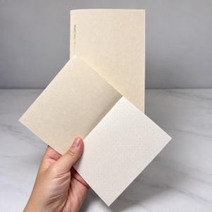 May include: A tall, cream-colored notebook with the text TRAVEL NOTE printed vertically in gold on the cover, paired with a smaller, open cream-colored notebook featuring a light gray dot grid pattern on the interior pages.