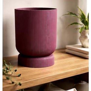 Puede incluir: Una maceta burdeos con estrías verticales, base redondeada y forma cilíndrica. Se encuentra sobre una estantería de madera. Una planta y libros son visibles en el fondo. Las líneas verticales crean un efecto texturizado.