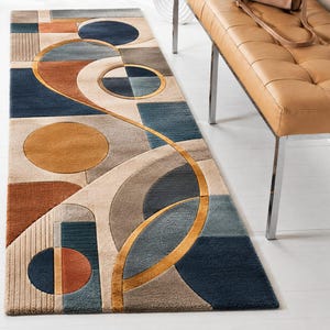 Op de afbeelding: Een rechthoekig vloerkleed met een abstract geometrisch patroon. Het ontwerp bevat cirkels en rechthoeken in tinten blauw, bruin, beige en goud. Het vloerkleed is waarschijnlijk gemaakt van een zacht materiaal, ideaal voor een woonkamer.