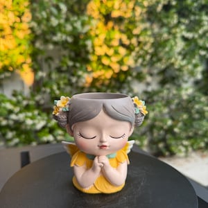 May include: A decorative planter shaped like a praying angel. The angel has gray hair, yellow and white flower accents, and a yellow dress. The planter is on a black surface with a blurred green and yellow background.