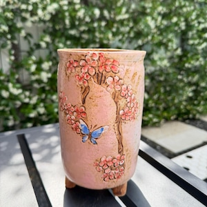 May include: A pink ceramic planter with a hand-painted design of a flowering tree and a blue butterfly. The planter has a textured surface and three small feet. The flowers are red and the leaves are brown.
